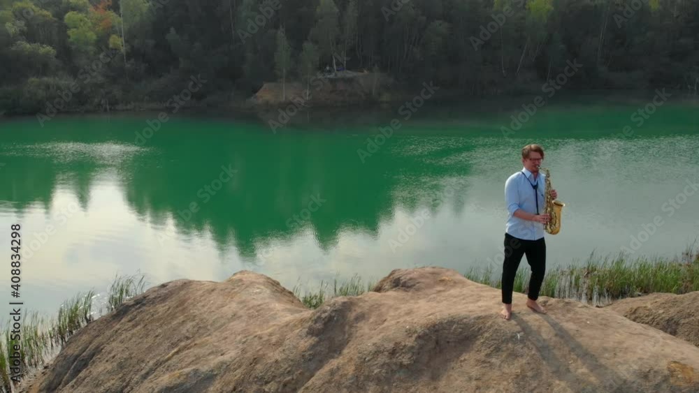 a young man with glasses and bare feet stands on a hill near a forest lake and plays the saxophone. the view from the top. aerial video recording a young man with glasses and bare feet stands on a hill near a forest lake and plays the saxophone. the view from the top. aerial video recording