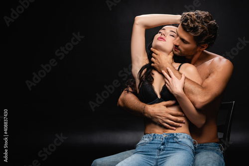 passionate young couple in jeans embracing with closed eyes while sitting on chair on black