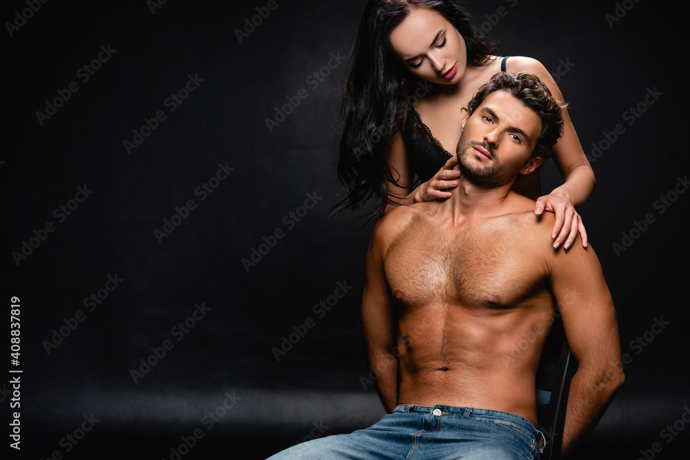 young muscular man looking at camera near seductive woman embracing him on black
