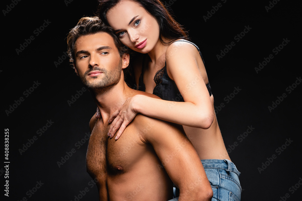 shirtless muscular man piggybacking seductive woman looking at camera isolated on black