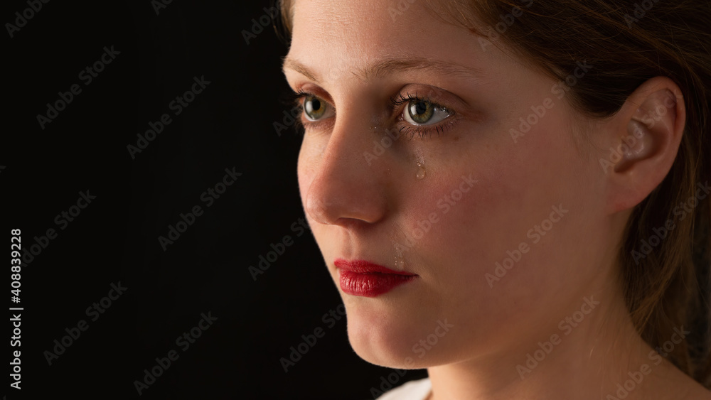 Fototapeta premium Sad woman crying on black background from close up. Unhappy girl with tears falling from eye on dark backdrop. Younge female feeling upset.