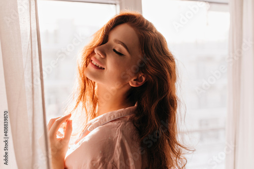 Indoor photo of sensual red-haired girl smiling with eyes closed. Pretty cherful woman posing near window. photo