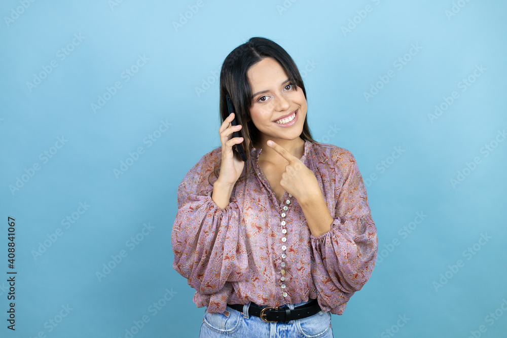 Beautiful latin woman wearing a pink shirt over blue isolated background having conversation talking on the smartphone smiling happy pointing with hand and finger Beautiful latin woman wearing a pink shirt over blue isolated background having conversation talking on the smartphone smiling happy pointing with hand and finger