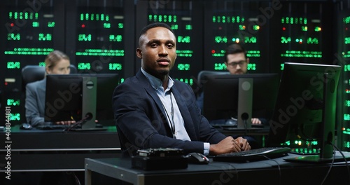 Smart African-American IT worker sitting in server room behind workplace working with database on computer. System software concept. photo