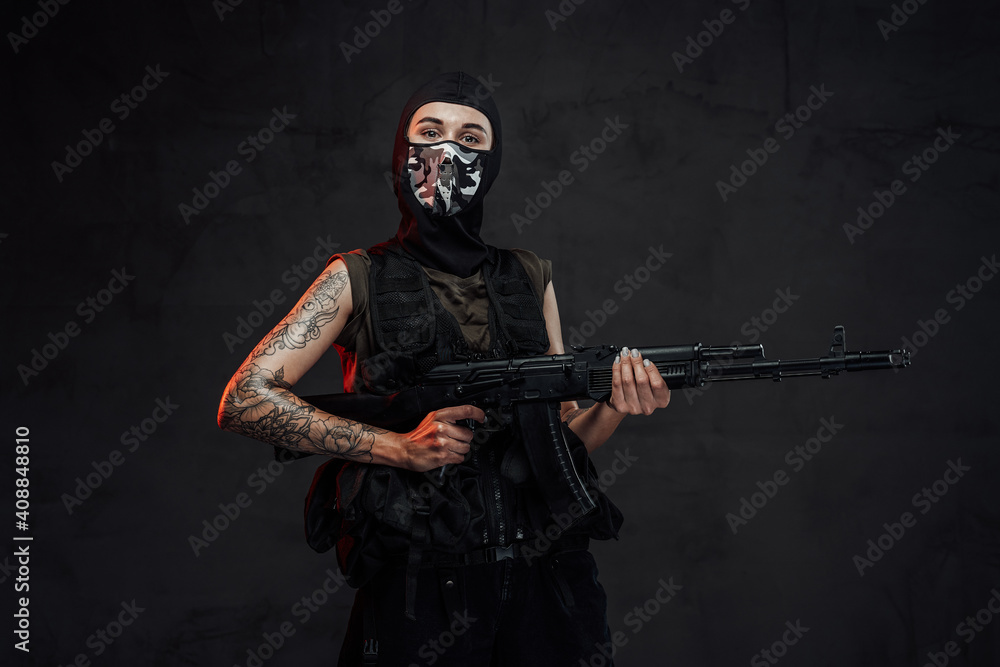 Tattooed and seductive woman in black armour and weared with mask poses in dark background holding assault rifle. Tattooed and seductive woman in black armour and weared with mask poses in dark background holding assault rifle.