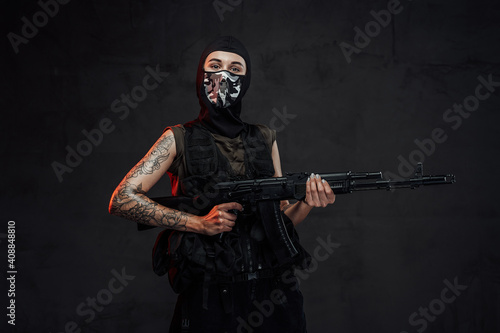 Fototapeta Naklejka Na Ścianę i Meble -  Tattooed and seductive woman in black armour and weared with mask poses in dark background holding assault rifle.