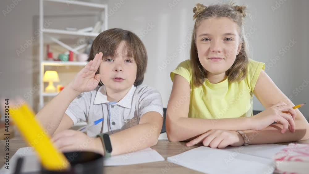 Brunette Caucasian boy raising hand and talking as girl sitting at desk looking at camera. Children studying online on lockdown. Wireless education concept. Brunette Caucasian boy raising hand and talking as girl sitting at desk looking at camera. Children studying online on lockdown. Wireless education concept.