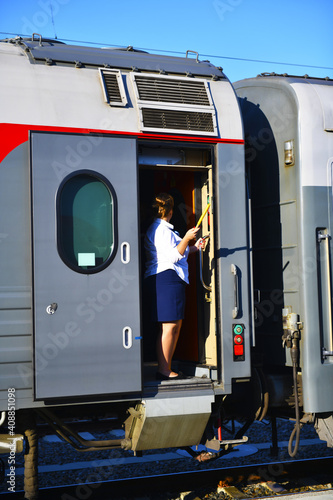 Railway cars. A female conductor stands in the doorway with a signal flag in her hand. Long-distance train, short stop at the railway station.