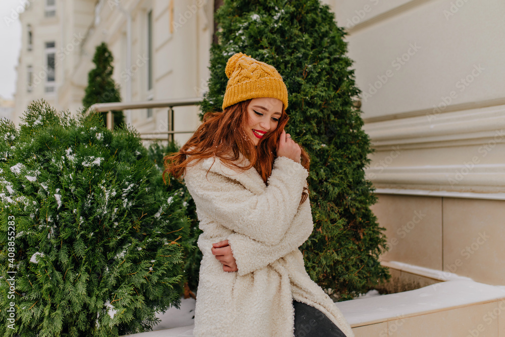 Ecstatic ginger girl posing near fir with sincere smile. Pleasant european woman in white coat standing outdoor.