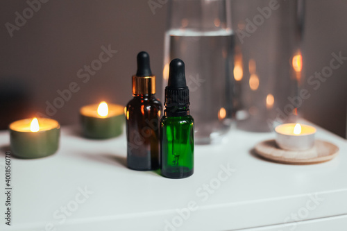 Two glass bottles of beauty oil on a table with candles, water, water glasses on a white table, at home