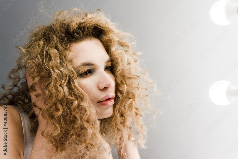 Mirror with bulbs reflection face of young curly blond woman