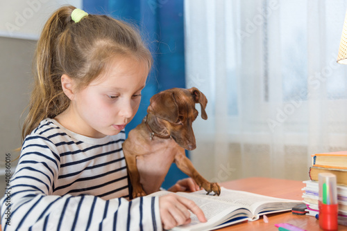 Child with dog reading book at home. Girl with pet sitting at window at read