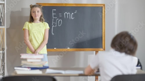 Confident intelligent Caucasian schoolgirl writing physical formulas on chalkboard in classroom and turning to camera. Blurred schoolboy sitting at desk at front. Children studying in school. photo