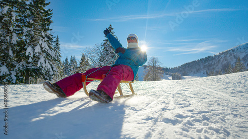 Fotografie LOW ANGLE Woman outstretches her arm while sledding down snowy hill on sunny day