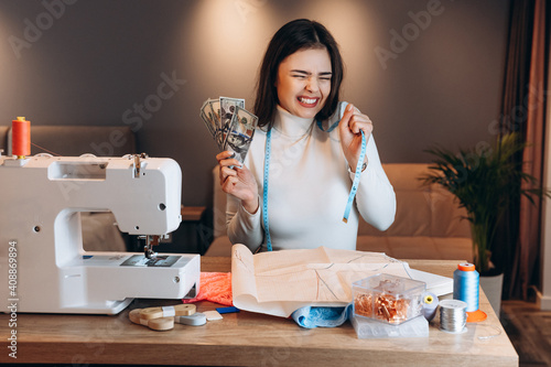Young dressmaker woman sews clothes on sewing machine. Looks happy holding tape measure and money in workshop. Creating online clothing design courses.