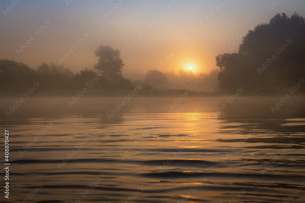 Obraz premium Spring nature landscape at dawn in foggy morning with bright sun on horizon. Amazing sunrise on river shore at mist. Natural scene of sun through fog. Scenery springtime