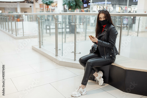 Girl with medical black mask and mobile phone in a shopping center. Coronavirus pandemic. A woman with a mask is standing in a shopping center. A girl in a protective mask is shopping at the mall