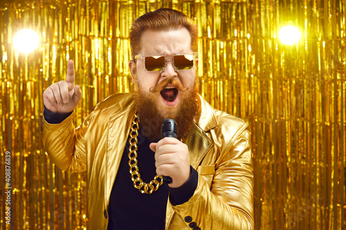 Funny singer in shiny golden jacket and gold chain holding microphone and singing songs at retro pop music concert. Happy bearded man performing at disco nightclub or enjoying time at karaoke party photo