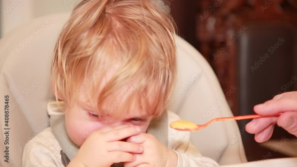 The baby refuses to eat delicious porridge. The child is naughty sitting in a child's chair in his kitchen. A caring father feeds his son with a spoon. Selective Focus The baby refuses to eat delicious porridge. The child is naughty sitting in a child's chair in his kitchen. A caring father feeds his son with a spoon. Selective Focus