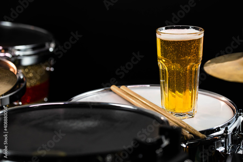 Glass of light beer on professional drum set closeup. Drumsticks, drums and cymbals, at live music rock concert, in the club stage, bar, or in recording studio. Black background.