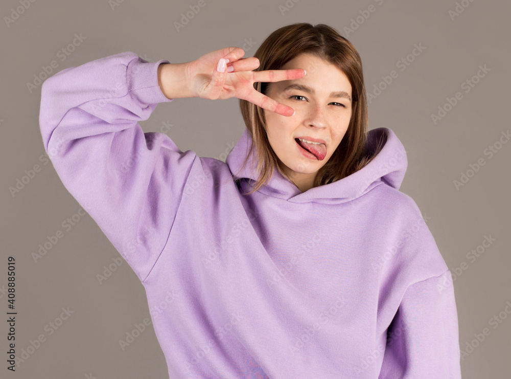 Portrait of happy young woman in purple sweater showing victory gesture by her fingers and playfully winking at the camera. Indoor studio shot isolated on gray background Portrait of happy young woman in purple sweater showing victory gesture by her fingers and playfully winking at the camera. Indoor studio shot isolated on gray background