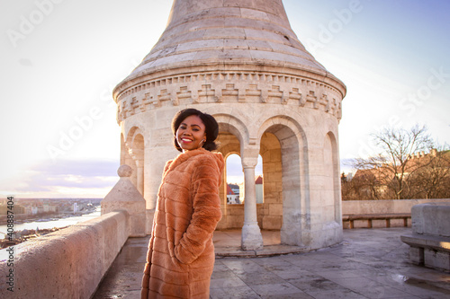 Black woman in an orange furry coat and red lips smilling joyfully at the light of a sunrise in front of view point