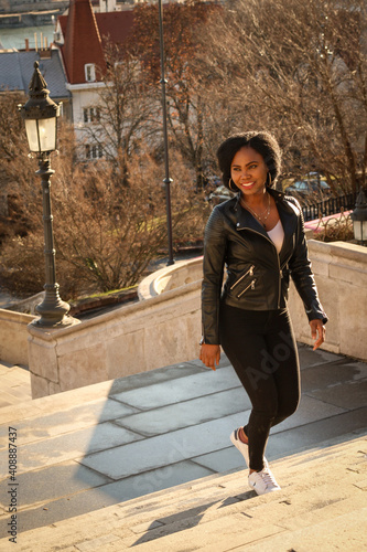 Dark skin girl in a black leather jacket and black pants walking up some stairs of yellow stone