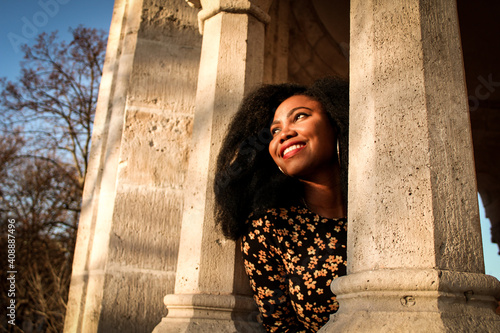 Black woman in a black dress with flowers and red lips smilling joyfully at the light of a sunrise between to columns made of stone