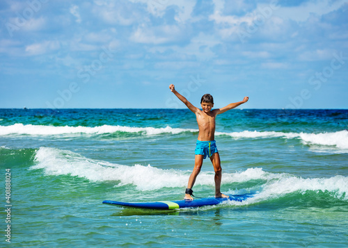 Young 10 years old boy posing with lifted hands ride surfboard on a wave in the sea © Sergey Novikov