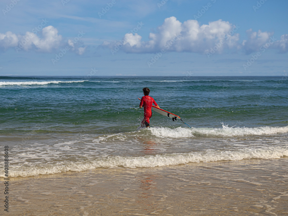 Deportista caminando dentro del agua vestido de rojo, con la tabla de surf en la Playa el Rosal en Cantabria, verano de 2020