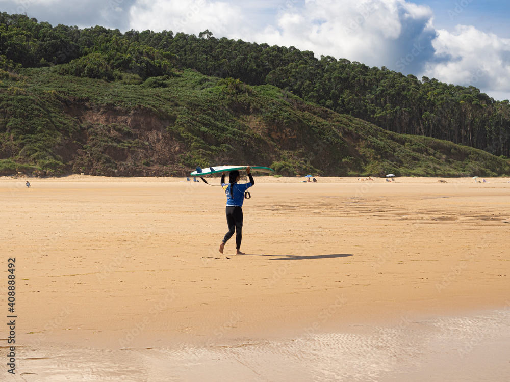 Chica deportista vestida de windsurfista andando con una tabla de surf sobre su cabeza, sobre la arena de n la Playa el Rosal en Cantabria, verano de 2020