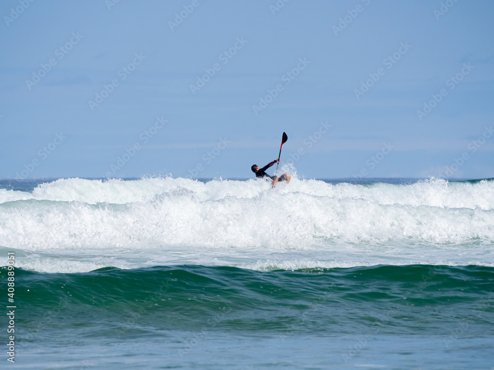 Padelsurfista cayendo de su tabla y con la pala al aire en el  mar Cantábrico sobre la espuma blanca de las olas de la Playa del Rosal en Cantabria, verano de 2020.