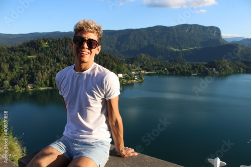 Latin skinny boy sitting in on a edge with a lake and montains behind wearing a white t-shirt, sunglasses, and blue swimming shorts, smiling
