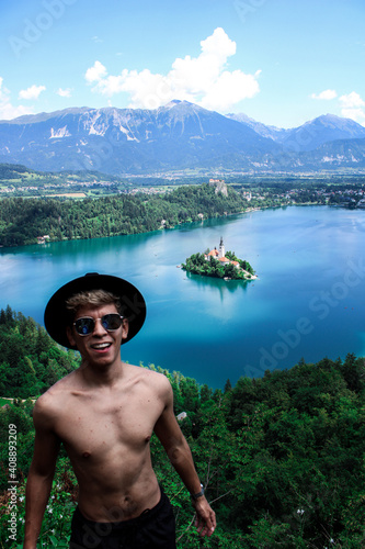 Latin skinny boy with dyed white hair in sunglasses with swimwear and a blak hat standing in a view point in a mountain with a blue lake and a island with a church in the middle
