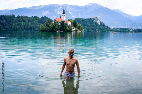 Latin skinny boy with dyed white hair in sunglasses in swimwear in a blue lake and a island with a church behind