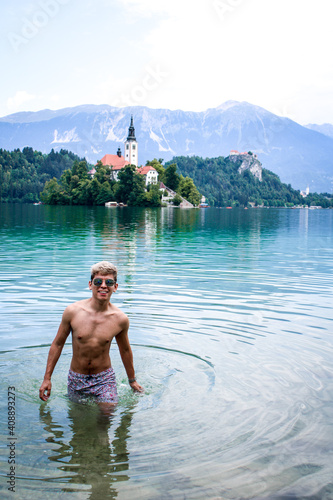 Latin skinny boy with dyed white hair in sunglasses and swimming shorts smiling in a blue lake and a island with a church behind