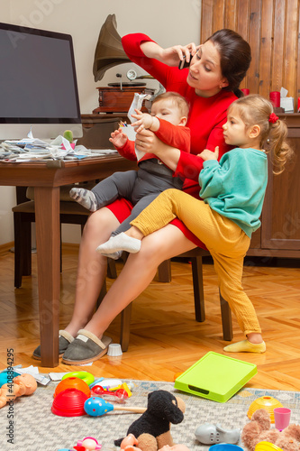 Mother with two child at home. Kids crying. Childcare and working mom. Woman talk by phone. Toddler tantrum. Young lady working at home during quarantine. Real people. A lot of bills on the table.