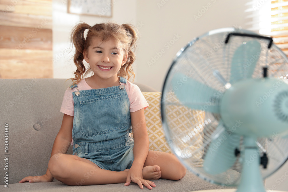 Little girl enjoying air flow from fan on sofa in living room. Summer heat - obrazy, fototapety, plakaty 