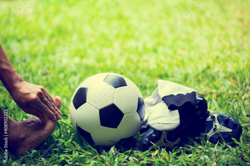 Soccer ball and sport shoes on green grass with soccer player in foot pain.