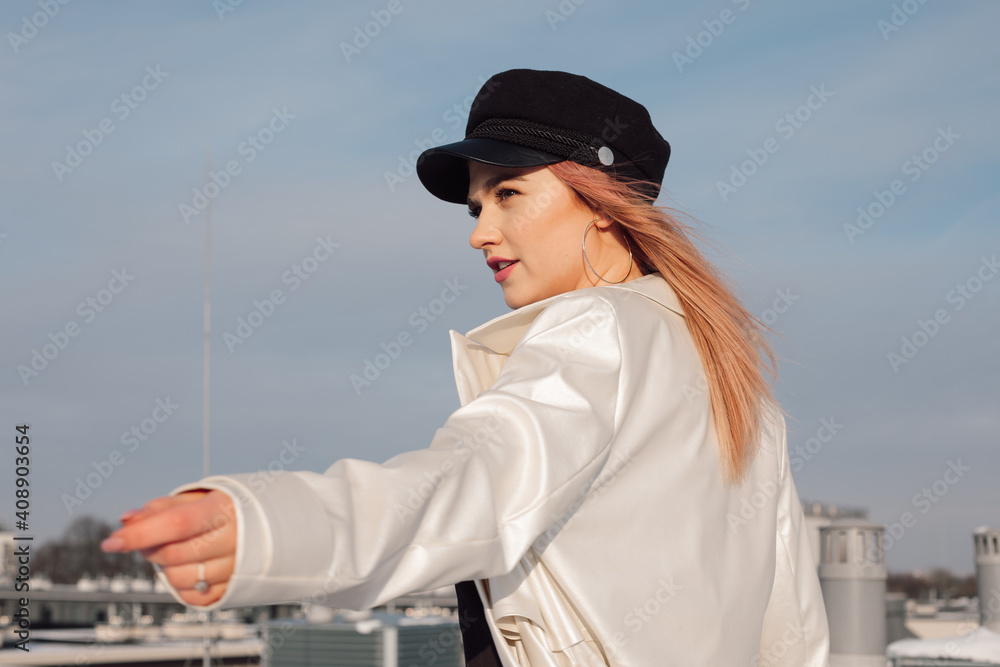 Girl against blue spring sky in light raincoat and cap on her head stretches out her hand in anticipation. Red-haired young beautiful woman looking to side. Close-up portrait
