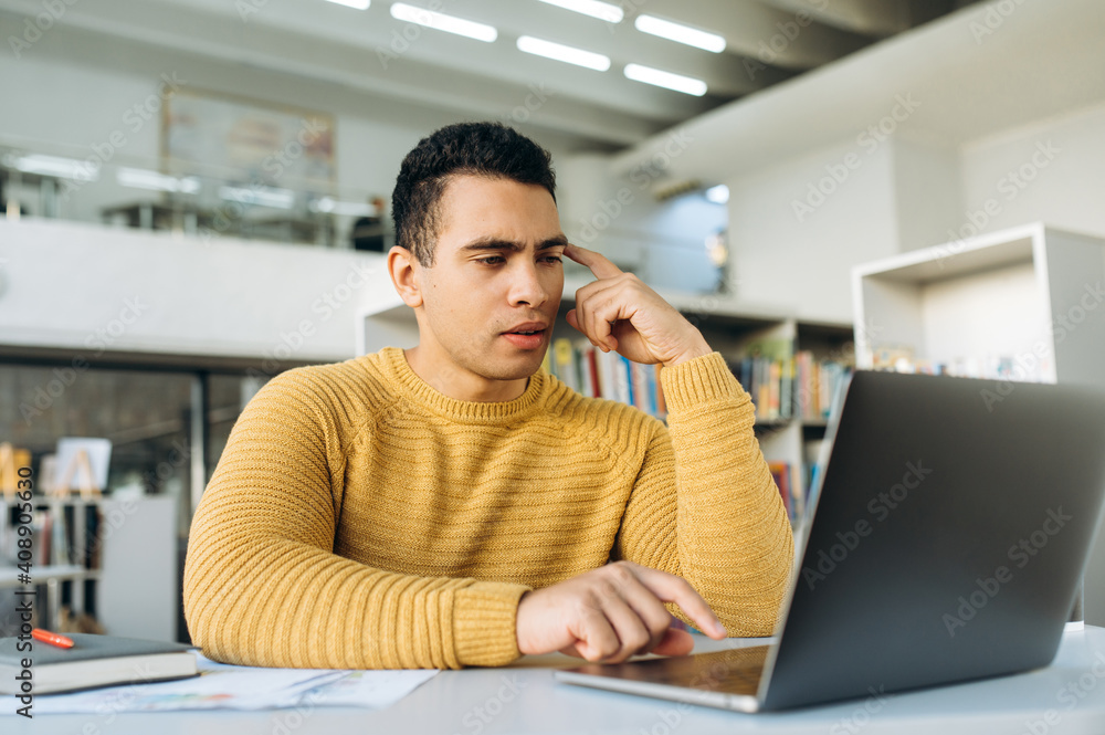 Serious hispanic guy sit at the work desk, using laptop. Concentrated male employee looking at the screen, browsing internet, search ideas, working on project Serious hispanic guy sit at the work desk, using laptop. Concentrated male employee looking at the screen, browsing internet, search ideas, working on project