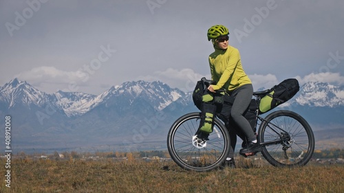 Fototapeta Naklejka Na Ścianę i Meble -  The woman travel on mixed terrain cycle touring with bikepacking. The traveler journey with bicycle bags. Sport tourism bikepacking, bike, sportswear in green black colors. Mountain snow capped.