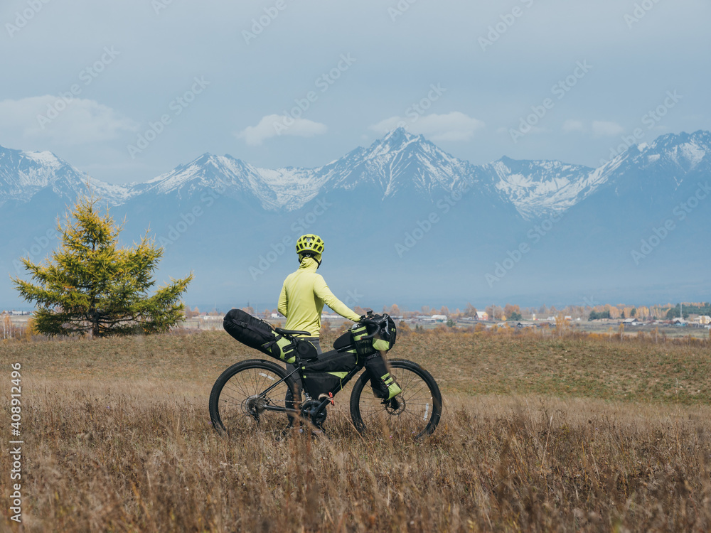Fototapeta premium The woman travel on mixed terrain cycle touring with bikepacking. The traveler journey with bicycle bags. Sport tourism bikepacking, bike, sportswear in green black colors. Mountain snow capped.