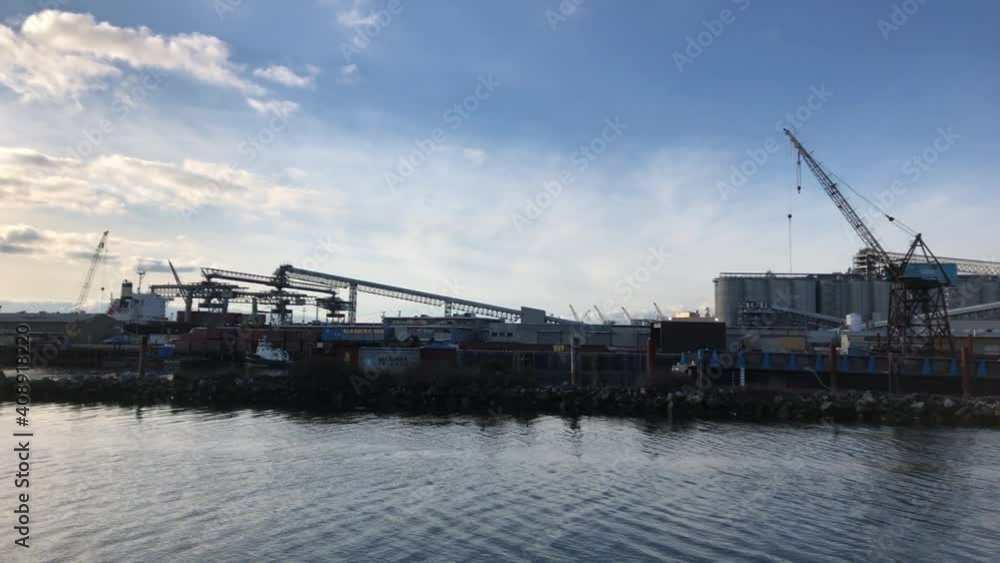 Multiple offshore vessels moored at a dock with on the background a big grain terminal with a bulk carrier alongside on a partly cloudy day while birds are flying around. Slowly panning shot