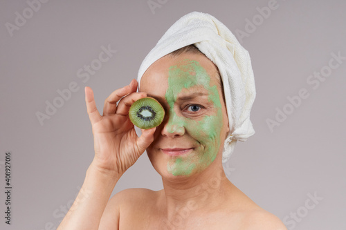 Mature woman with bare shoulders, white towel on her head, applied green fruit mask on her face, kiwi closed her eyes. Facial skin care concept. Studio shot over gray background.