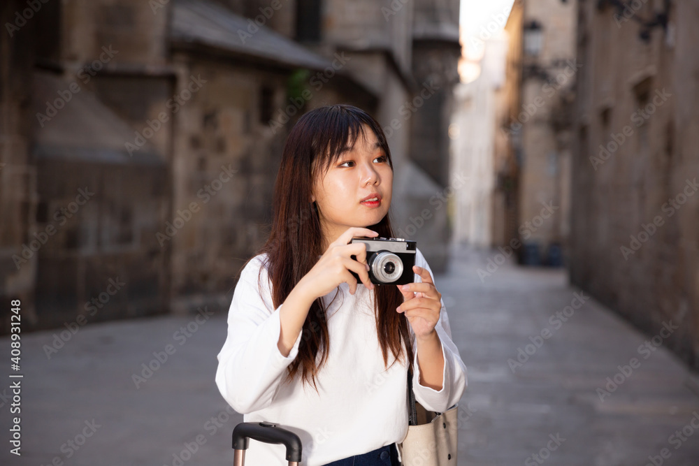 Young chinese woman traveler strolling with luggage around city, making photo of sights. High quality photo