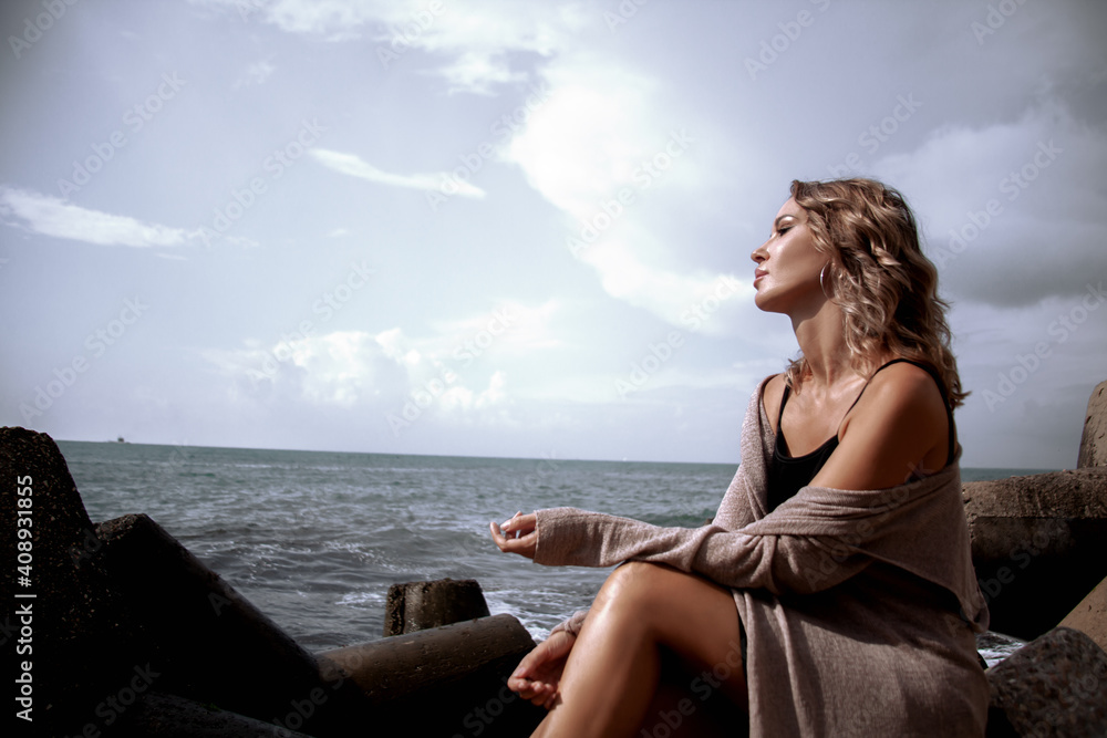 Beautiful sexy woman sitting by the sea with dramatic stormy sky , enjoys the wind and look. Alone woman dressed black dress and beige cardigan