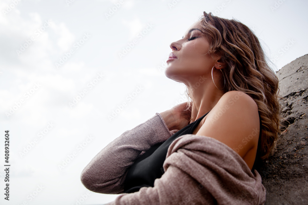 Beautiful sexy woman sitting by the sea with dramatic stormy sky , enjoys the wind and look. Alone woman dressed black dress and beige cardigan Beautiful sexy woman sitting by the sea with dramatic stormy sky , enjoys the wind and look. Alone woman dressed black dress and beige cardigan