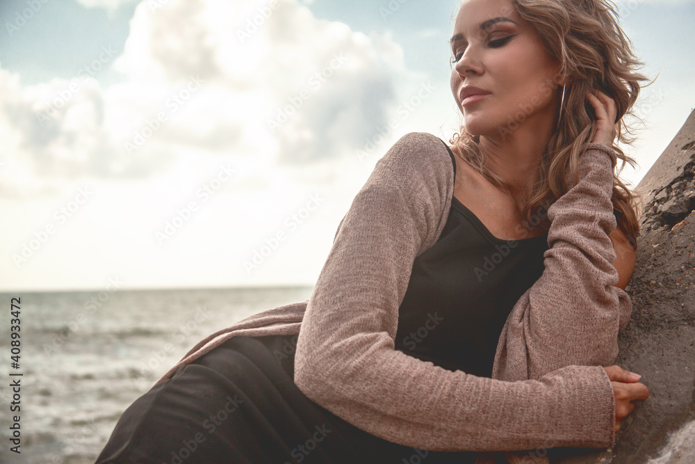 Portrait of beautifukl sexy woman with smoky-eyes make-up sitting by the sea with dramatic stormy sky , enjoys the wind and look. Alone woman dressed black dress and beige cardigan