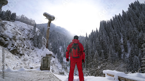 The guy stands on the edge of the hill and enjoys the view. Looks at a snowy gorge with high mountains, and fir trees growing on the cliffs. Clouds cover the mountain. Mountain suit with backpack. photo